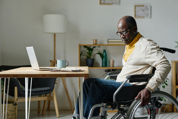 African mature man sitting on wheelchair at table with laptop, he spending time at home during his...