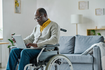 African mature man with disability sitting in wheelchair and communicating online using his laptop...