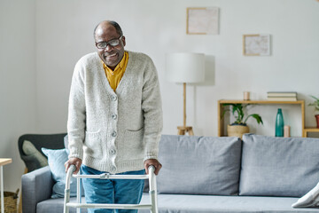 Portrait of African senior man with disability smiling at camera while learning to walk with walker in the room - obrazy, fototapety, plakaty