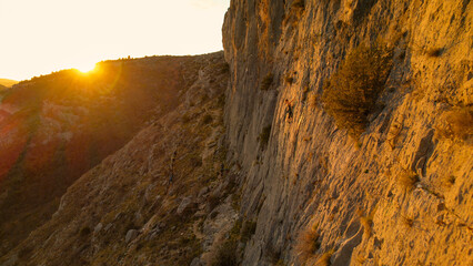 AERIAL: Female climber ascending up the wall with golden sun flare in background