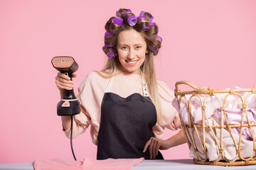 A smiling woman with curled rollers in her hair holds a steamer to iron shirts. Portrait of a beautiful happy girl on a pink background.