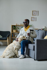 African senior man in dark glasses with stick sitting on sofa with guide dog in the living room