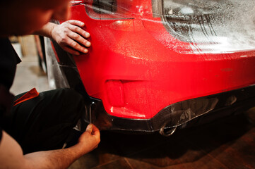 Car service worker put anti gravel film on a red car body at the detailing vehicle workshop. Car protection with special films.