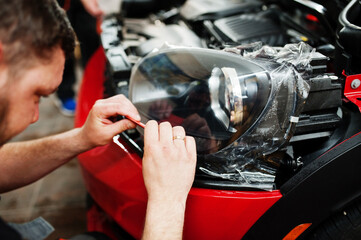 Car service worker put anti gravel film on a red car body at the detailing vehicle workshop. Car protection with special films.