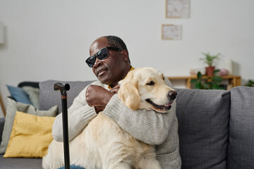 African senior man in glasses with bad sight embracing his individual guide dog while sitting on...