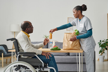 African young caregiver unpacking shopping bag with food she delivering for man with disability