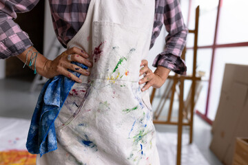 Image of midsection of biracial female artist in painted apron in studio