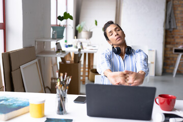 Image of tired biracial female artist using laptop and stretching in studio