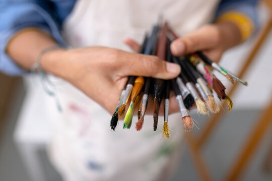 Image of hands of biracial female artist with brushes in studio