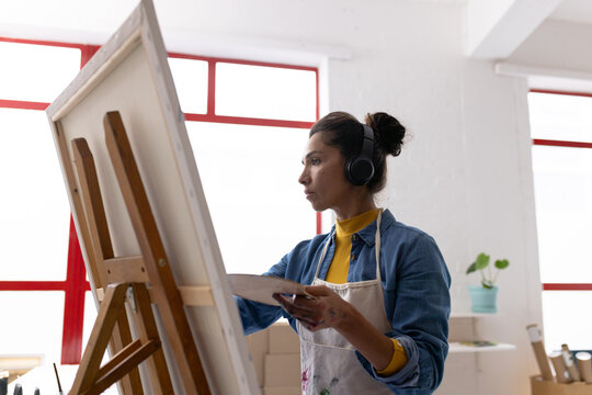 Image of back view of biracial female artist in headphones working on painting in studio