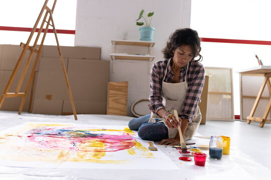 Image of biracial female artist painting on floor in studio