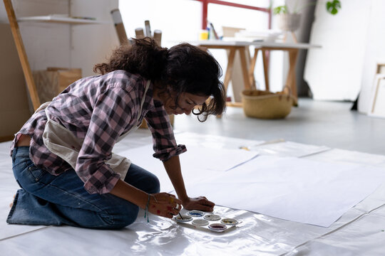 Image of biracial female artist painting on floor in studio