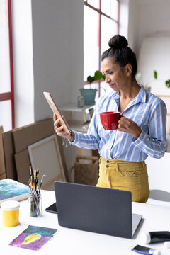 Vertical image of biracial female artist holding painting and drinking coffee in studio
