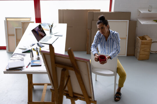 Image of thoughtful biracial female artist sitting on chair and drinking coffee in studio