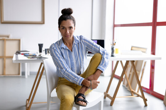 Image of thoughtful biracial female artist sitting on chair in studio
