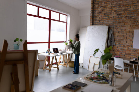 Image of happy biracial female artist using laptop and smartphone and working in studio