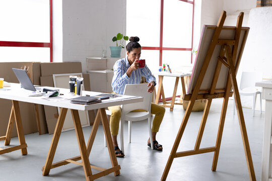 Image of thoughtful biracial female artist sitting on chair and drinking coffee in studio