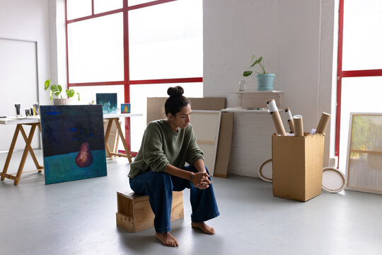 Image of thoughtful biracial female artist sitting in studio