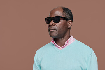 Portrait of African senior blind man in dark glasses standing against brown background