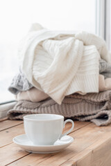 A white cup stands on the windowsill, a pile of warm clothes in the background