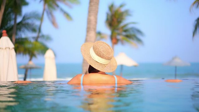 Back View of Unrecognizable Woman With Floppy Hat and Bikini Standing in Infinity Swimming Pool and Looking at Tropical Beach and Sea Horizon