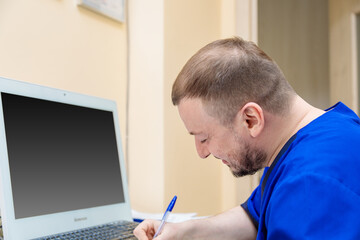 A male ophthalmologist sits at a workplace at a computer and writes with a pen