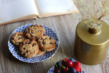 Plate of chocolate chip cookies, cup filled with strawberries, blueberries and cherries, open book and vase with gypsophila flowers on the table. Selective focus.
