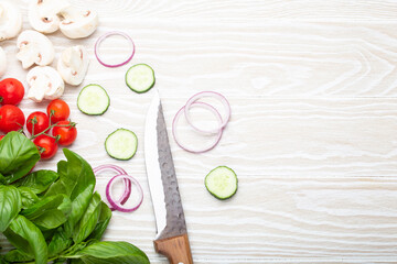 Obraz na płótnie Canvas Food cooking background: fresh ingredients with kitchen knife, basil, cherry tomatoes, mushrooms, cut cucumber and onion on white wooden background top view. Meal preparing template, space for text