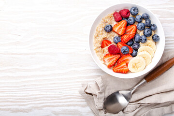 Oatmeal porridge with fruit and berries in bowl with spoon on white wooden background table top view, homemade healthy breakfast cereal with strawberry, banana, blueberry, raspberry. Space for text