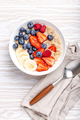 Oatmeal porridge with fruit and berries in bowl with spoon on white wooden background table top view, homemade healthy breakfast cereal with strawberry, banana, blueberry, raspberry