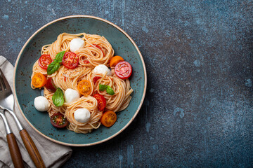 Delicious spaghetti with mozzarella, colourful cherry tomatoes, fresh basil on ceramic plate on rustic stone background top view. Italian healthy tasty food pasta for dinner or lunch. Space for text