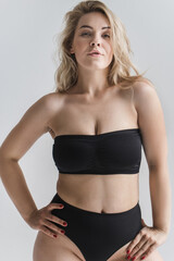 Sensual blonde plus size languidly posing on a gray background in black sports underwear. The woman bit her lips. Chubby girl in black lingerie