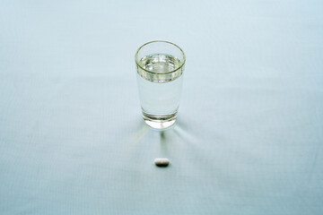 White capsule-shaped tablet in front of a transparent glass of water on a light surface. The...