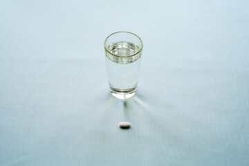 White capsule-shaped tablet in front of a transparent glass of water on a light surface. The concept of one dose, taking the medicine, control, moderation, caution, calculation. High angle.