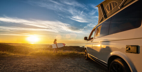 Surfer girl sitting near her mini van and looking on the ocean at summer sunset  with a surfboard...