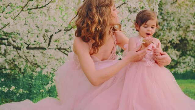 Happy cute woman mother tenderly hugs daughter. Little girl opened mouth. Princess holds in hand sprig spring cherry tree blooming white flowers. Same long pink ball gowns. Concept maternal happiness
