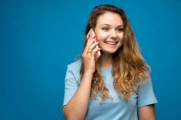 Fototapeta premium Young smiling happy fun woman in blue basic casual t-shirt talking speak on mobile phone conducting pleasant conversation on blue background studio portrait.