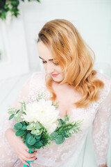 Obraz na płótnie Canvas Young attractive bride with blond hair with wedding makeup and hairstyle in a white lace peignoir with bride's bouquet in a bright interior