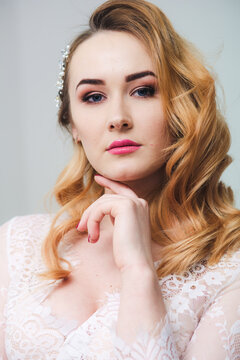 Young attractive bride with blond hair with wedding makeup and hairstyle in a white lace peignoir in a bright interior