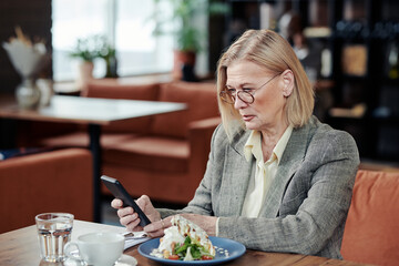 Mature businesswoman in suit using mobile phone while having lunch at the table in cafe