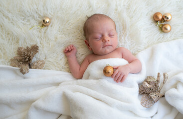 Baby holds golden ball in hand and soundly sleeps on white blanket decorate by golden balls and branches Christmas tree. Newborn is happiness for family. Top view. New year