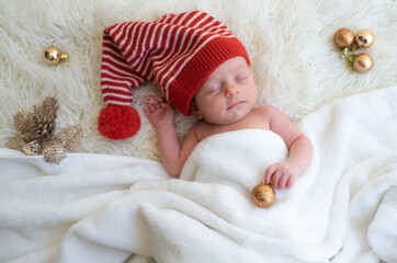 Baby in Santa hat and with golden ball in hand sleeping soundly and peacefully on white blanket. Newborn is happiness for family. Happy new year and Christmas. funny elf and fairy. Top view.