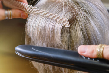 Professional hairdresser drying hair with a hair straightener client. Woman at the hairdresser. Shallow depth of field. Focus on a hairbrush and hair in a center frame