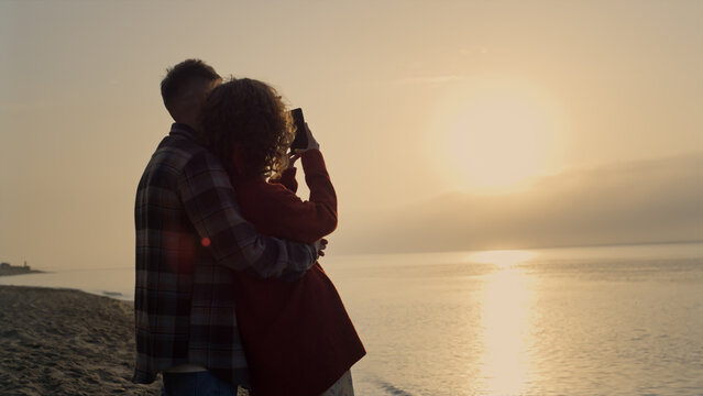 Man embracing woman on beach at sunrise. Girl making photo of sea landscape