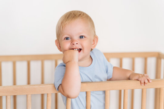 Indoor Portrait Of Happy Blond Baby Boy On White Background Growing First Milk Tooth Standing In Wooden Bed With Hand In Mouth Massaging Itchy Gums. Teething Concept