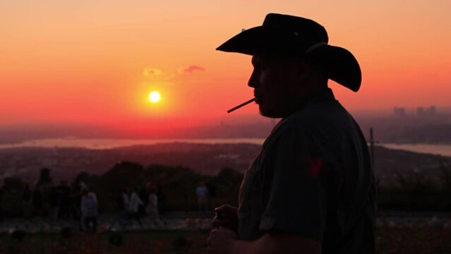 Silhouette man in a hat smoking a small cigar at an orange sunset in the evening