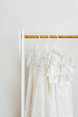 White summer children's dresses with flowers on hangers in wardrobe. Wooden Clothing Rack with children's outfits. Nursery Storage Ideas. Home kids wardrobe.