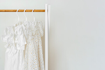 White summer children's dresses with flowers on hangers in wardrobe. Wooden Clothing Rack with children's outfits. Nursery Storage Ideas. Home kids wardrobe.