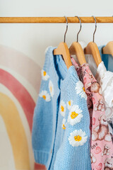 Wooden Clothing Rack with children's outfits. Blue knitting jacket, blouse, dress and sweaters on hangers in wardrobe close up. Nursery Storage Ideas. Home kids wardrobe.
