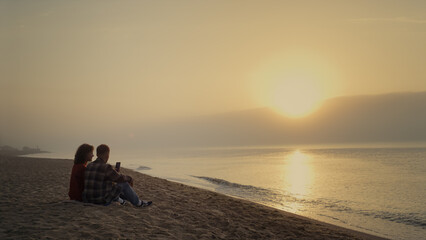 Couple sitting on beach at sunset. Woman and man enjoying ocean landscape 